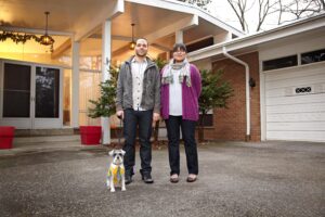 A man and a woman standing in front of a home with their pet dog.