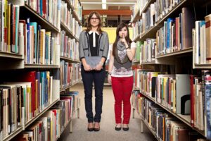 Two women standing in a row of books at the library. One of them is holding their books and another is shushing at the camera.